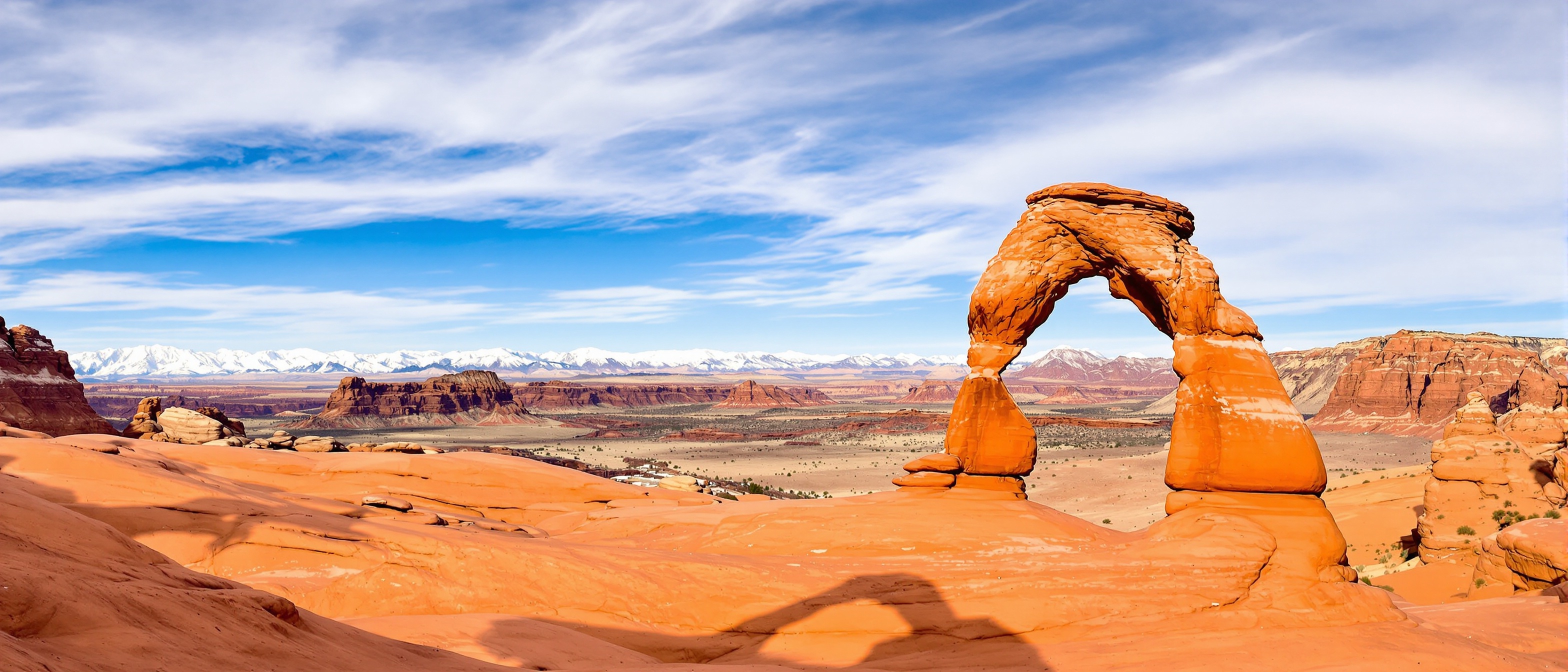 Stunning panoramic view of Delicate Arch in Arches National Park Moab Utah with dramatic red sandstone natural arch formation standing majestically against brilliant blue sky vast desert landscape stretching to distant snow capped La Sal Mountains on horizon golden afternoon sunlight illuminating vibrant orange and red rock surfaces rugged slickrock terrain in foreground few scattered desert plants clear sky with wispy clouds iconic Southwest geological formation natural wonder epic scale and beauty
