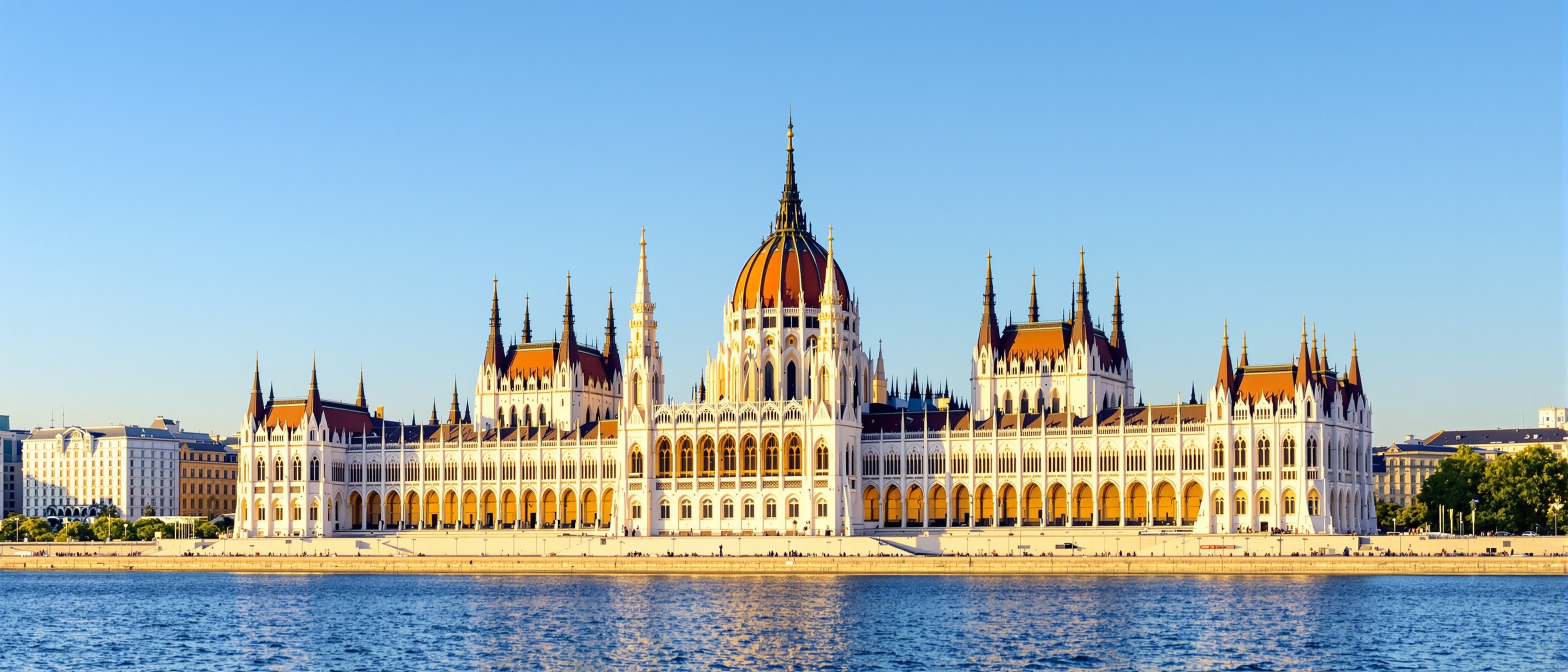 Magnificent Hungarian Parliament Building in Budapest viewed from across Danube River, stunning Gothic Revival architecture with ornate spires and central dome, golden afternoon light illuminating cream colored facade, intricate architectural details and decorative elements, calm river waters reflecting building, iconic landmark along riverbank